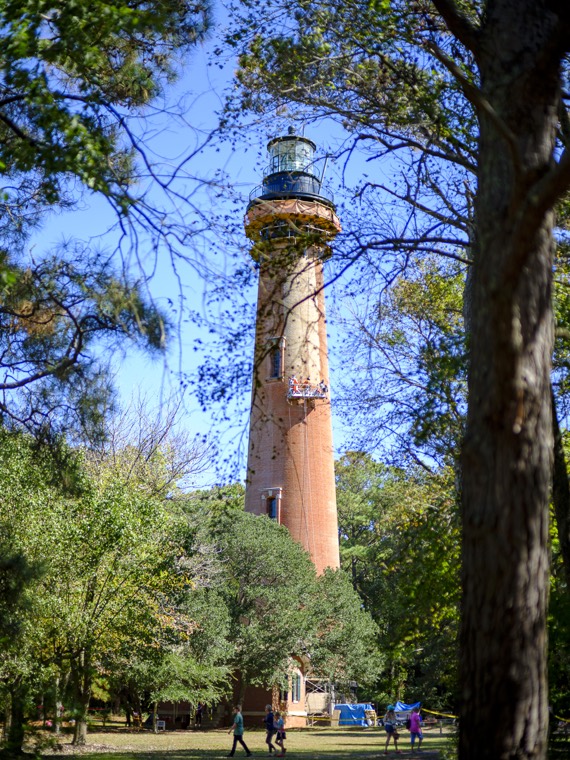 The red-brick Currituck Beach Lighthouse rising above green trees, with visitors at its base.