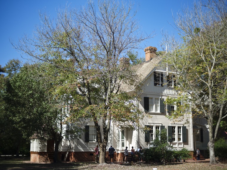 A large white house with green shutters shaded by trees, a few people sitting near the porch.