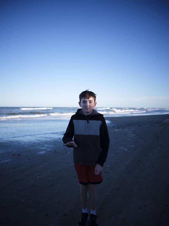 A kid in a hoodie and red shorts holding up a small find on the beach, waves behind.