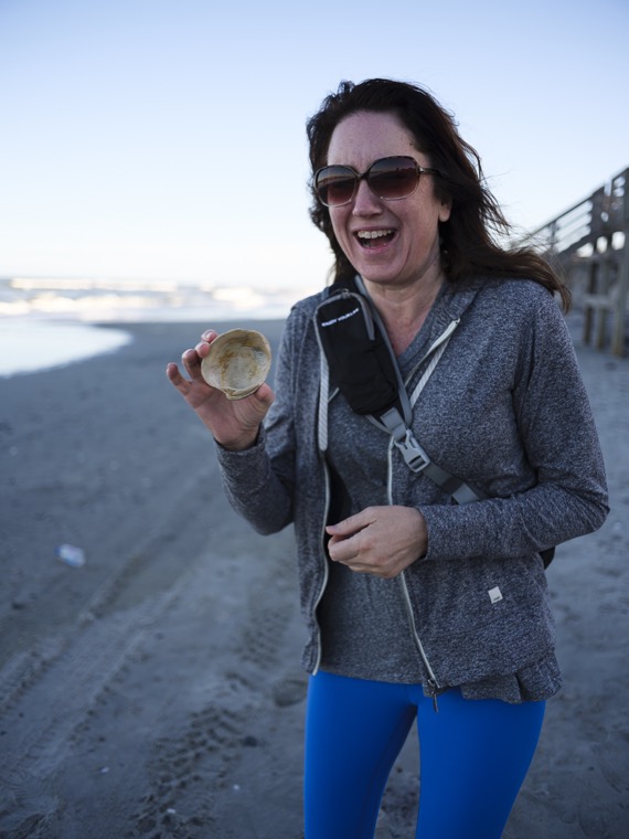 A laughing person holding up a shell for the camera, in a grey hoodie and blue leggings, with a wooden pier in the background.