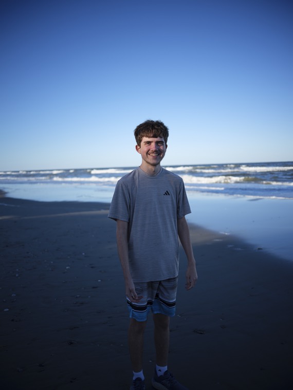 A smiling teenager in a grey t-shirt standing on the beach with waves rolling in behind.