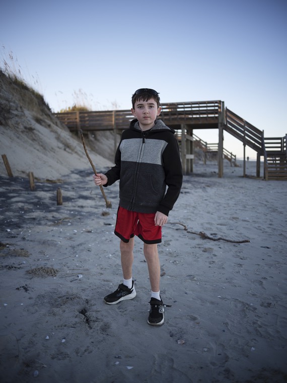 A kid holding a long stick on the sand, with dunes and a wooden beach walkway behind.