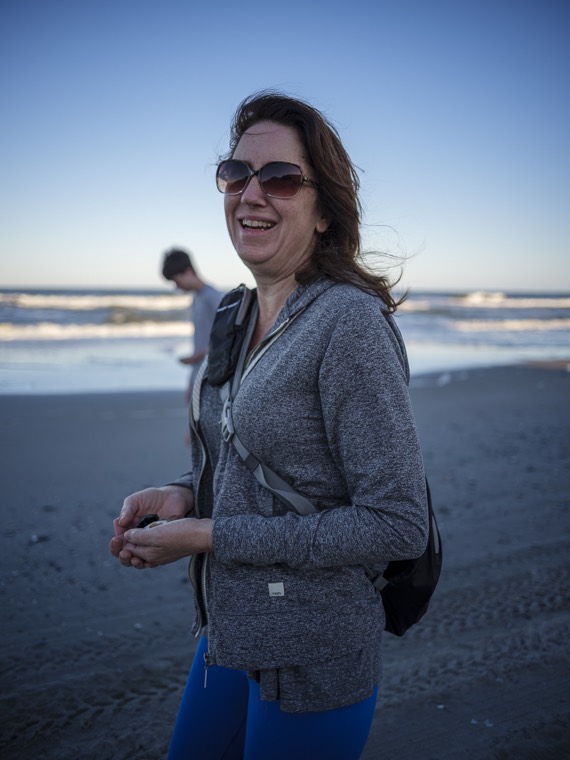 A person laughing with hair blowing in the wind, someone standing near the surf in the background.